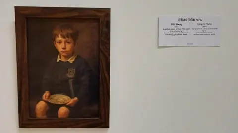 BBC A brown frame that has an image of a school boy holding a round plate on a white wall. There is a white piece of paper next to it that has Elias Marrow and Empty Plate 2024 written on it.