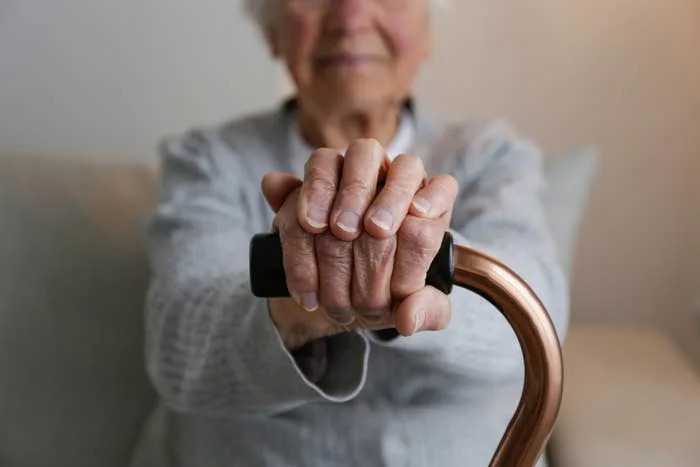 Older adult sitting with hands resting on a cane, wearing a light shirt, conveying a sense of calmness and stability