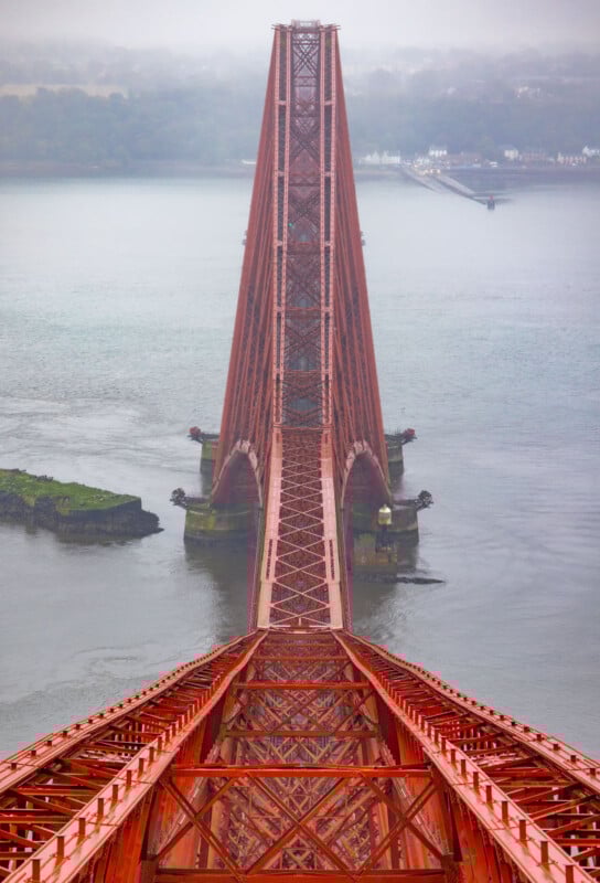 A view from above the central span of a red steel bridge crossing a wide river, with misty hills and buildings visible in the background.