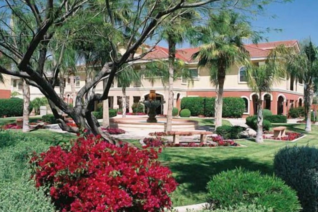 A resort with palm trees, green grass, red flowers, and a large fountain in front of it.