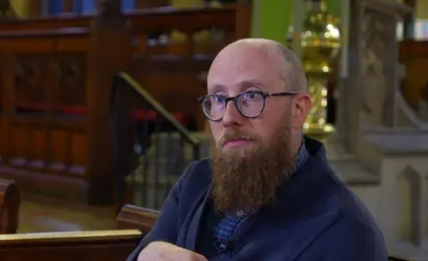 A mostly bald man wearing glasses and with a brown long bears, sits in front of church furniture while wearing a navy blue jacket. He is straight-faced, showing little emotion.