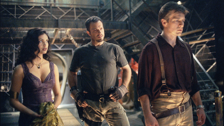 Inara Serra (Morena Baccarin) and Jayne Cobb (Adam Baldwin) stand behind Mal Reynolds (Nathan Fillion) in a cargo hold in Firefly