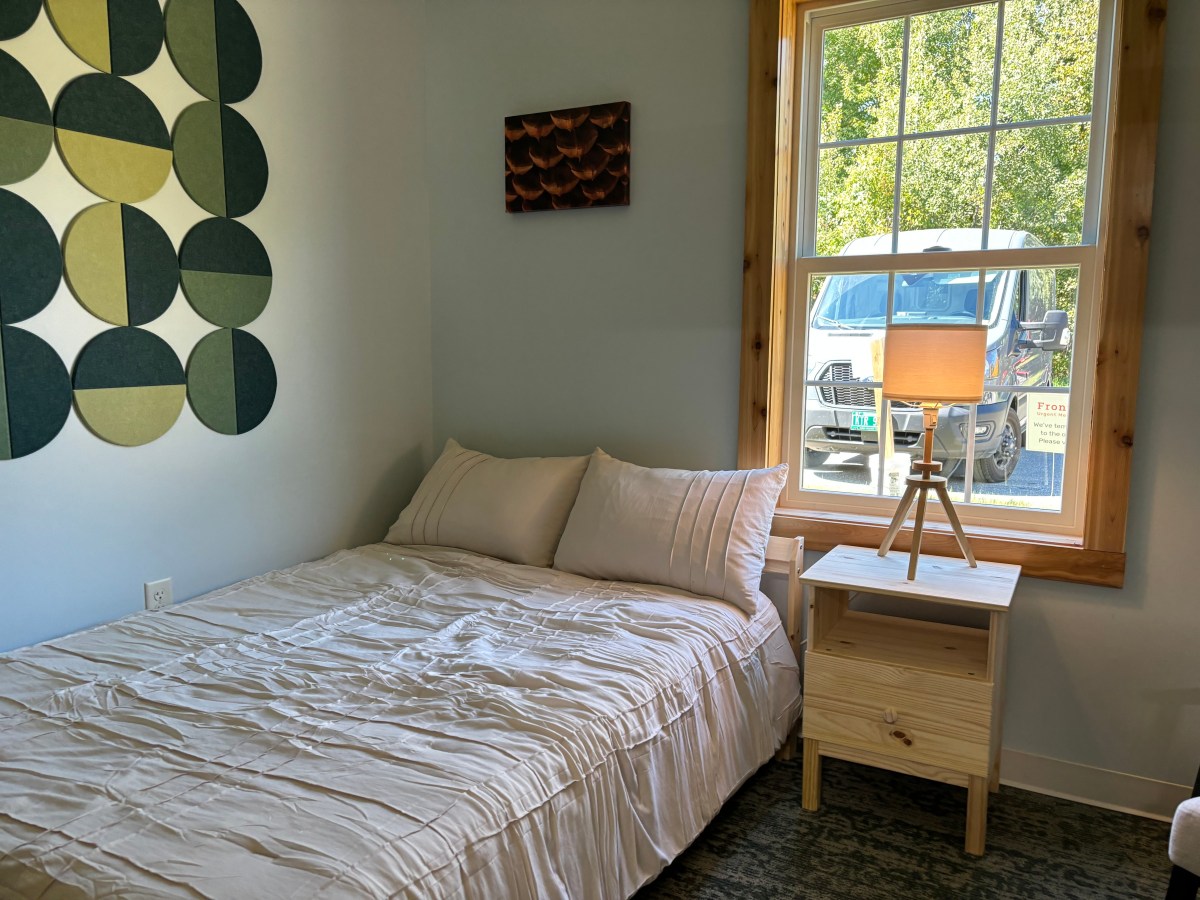 A neatly made bed with white bedding beside a wooden nightstand and lamp, next to a window with a view of a parked van and green trees outside.