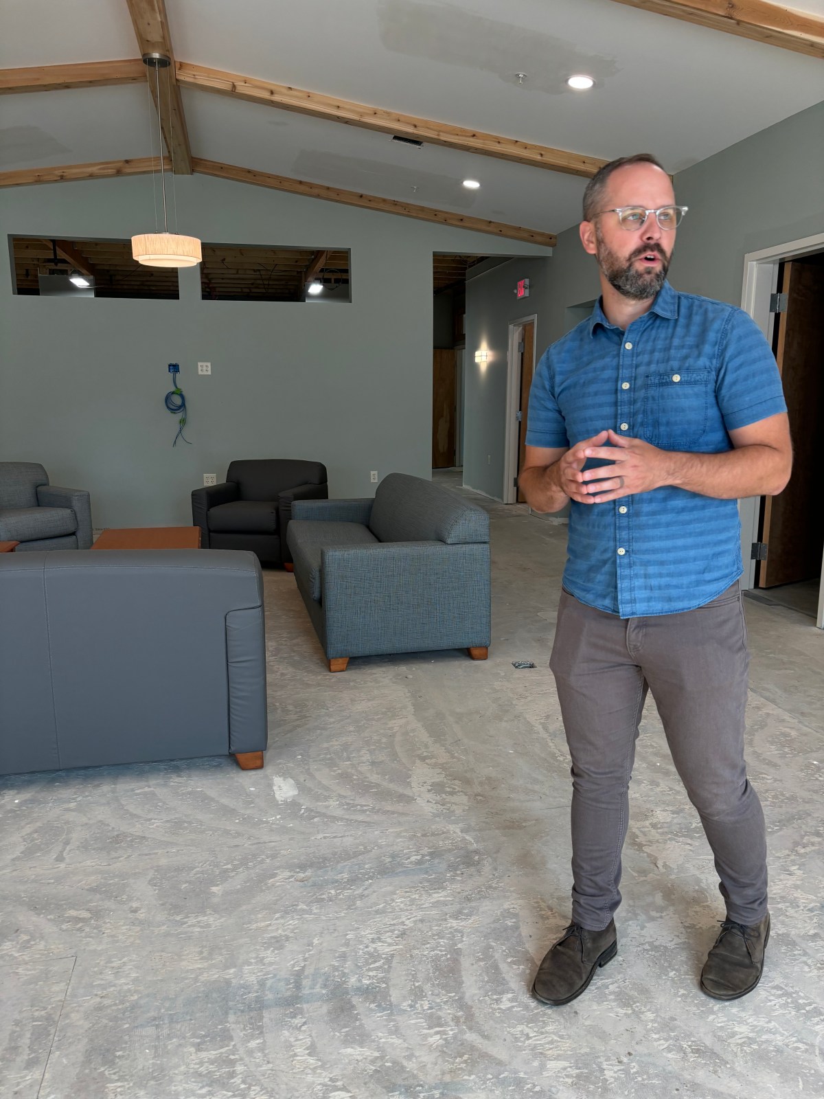 A man in glasses and a blue shirt stands in a sparsely furnished room with multiple couches and unfinished flooring.