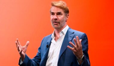 Erik Brynjolfsson gestures while speaking onstage during a panel, with a vivid orange background behind him.