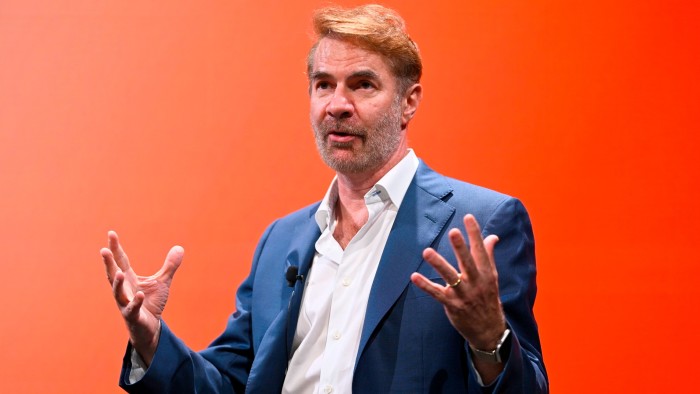 Erik Brynjolfsson gestures while speaking onstage during a panel, with a vivid orange background behind him.
