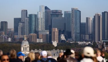 Tourists stand at a viewpoint with the Canary Wharf skyline and skyscrapers in the background on a clear day.