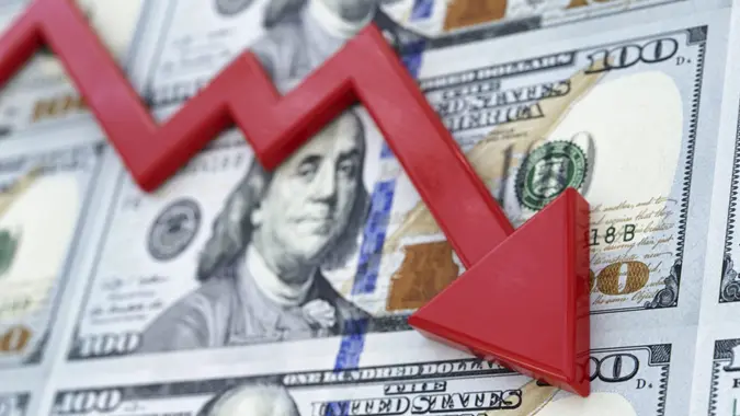 A few one hundred dollar bills with a red arrow on top of it going downward, symbolizing the US economy crashing.