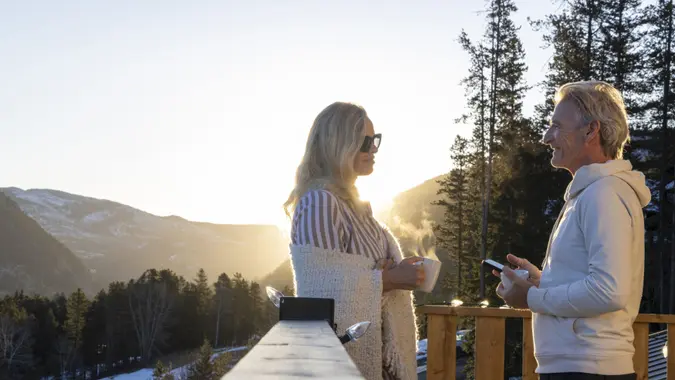Mature couple chat on chalet deck at sunrise in winter, with phone and hot drink, Canadian Rockies.