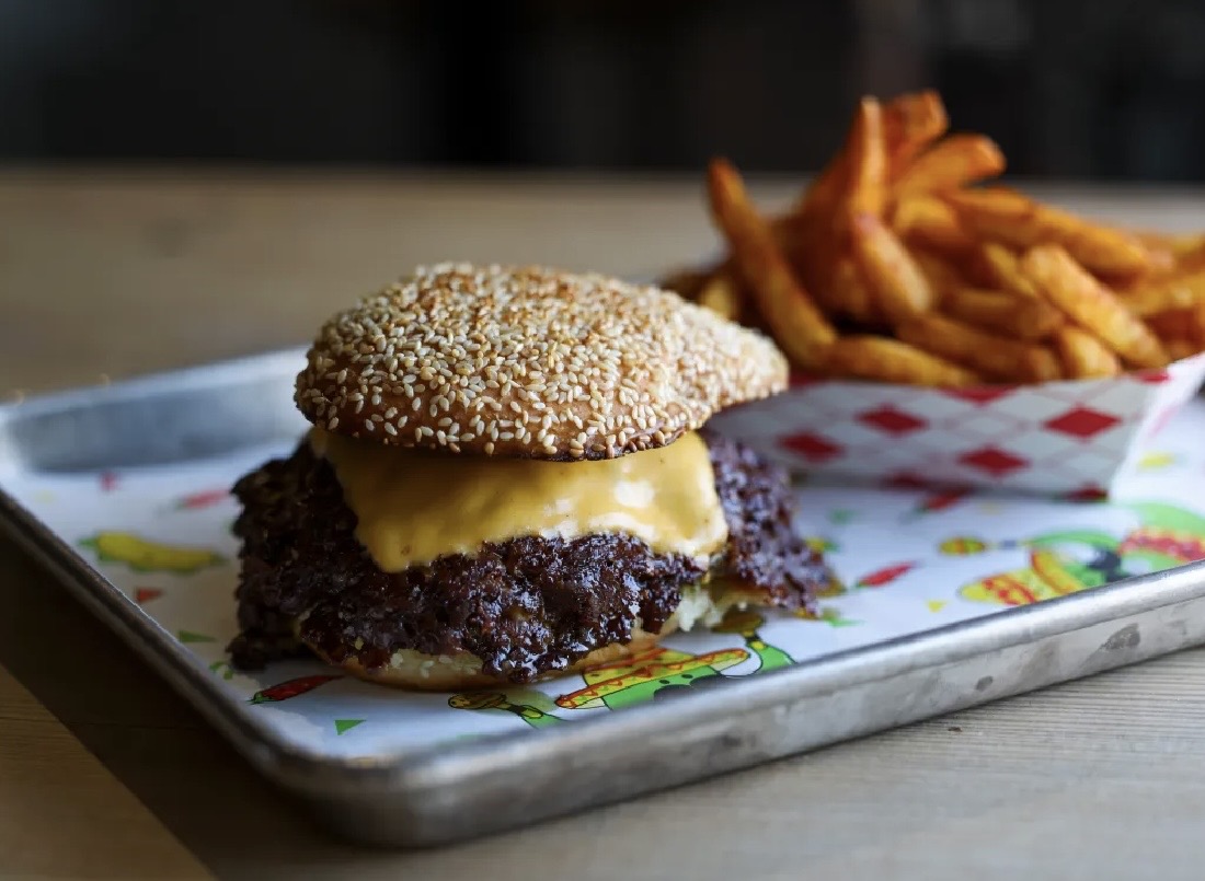 A lacy smashburger on a thin, toasted poppyseed bun with melted cheese on a metal tray with a side of fries in a paper boat.