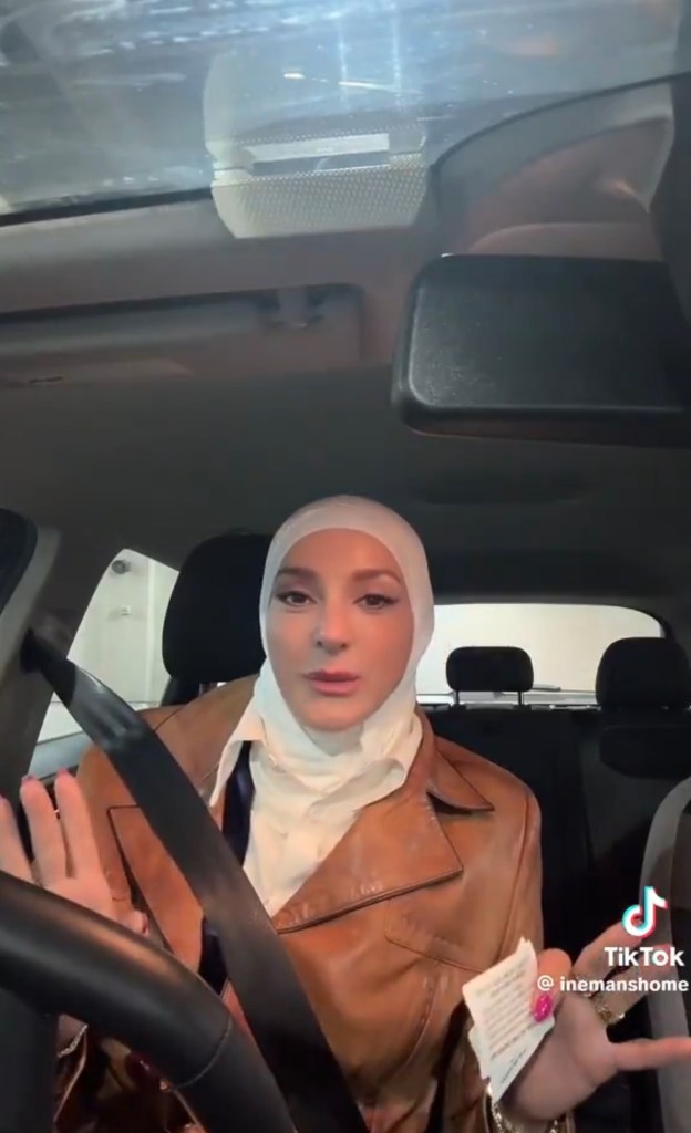 Iman Masoud, wearing a white hijab and brown jacket, in a car, speaking with her hands while holding a small piece of paper.
