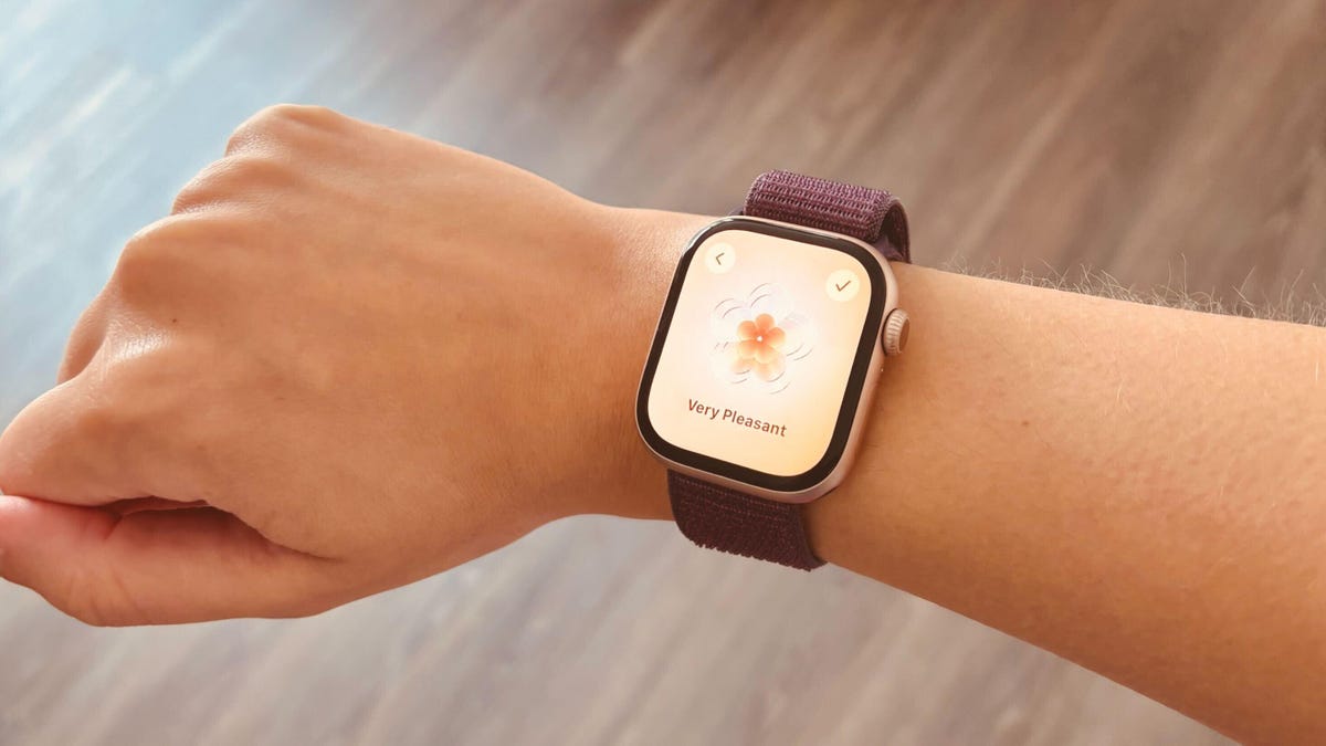 A hand with an Apple Watch 10 on it showing the State of Mind feature over a wood floor.