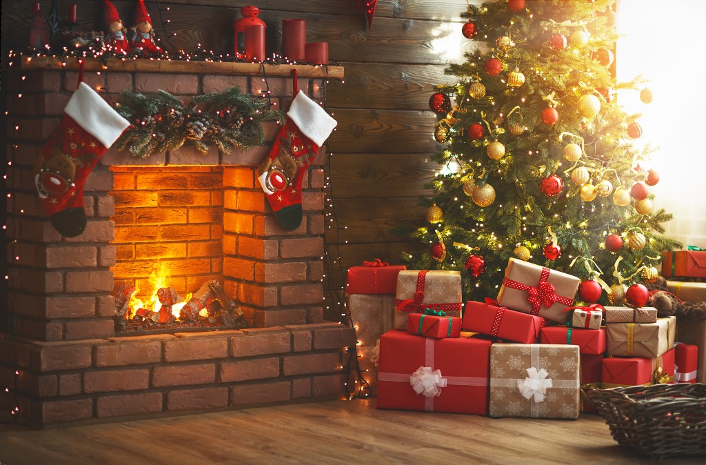 A decorated Christmas tree with gifts by a lit fireplace with stockings.