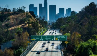 Cars drive along a freeway lined with green hills toward downtown Los Angeles, with tall skyscrapers visible in the background and green highway signs overhead directing traffic.