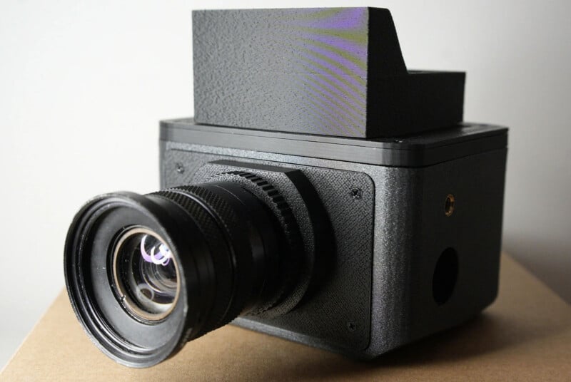 A close-up view of a black, boxy camera with a large lens and a rectangular block on top, placed on a brown surface against a light background.