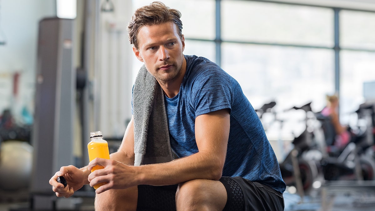Handsome young man in sportswear holding bottle of fresh orange juice while resting at gym, looking thoughtful and serious while taking a break after fitness exercise on bench.