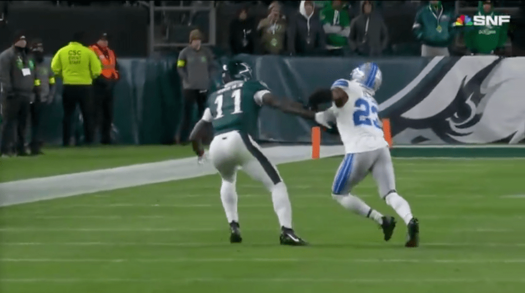 Two American football players, one in white and green, the other in white and blue, are on a green field.