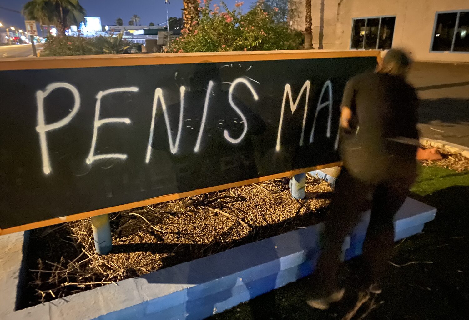 A male dressed in black clothing spray paints the words "PENIS MAN" on a sign.
