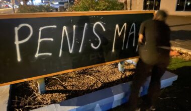 A male dressed in black clothing spray paints the words "PENIS MAN" on a sign.