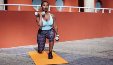 A person wearing a light blue exercise top and purple patterned leggings lifting light blue weights on a bright yellow yoga mat outside on concrete.