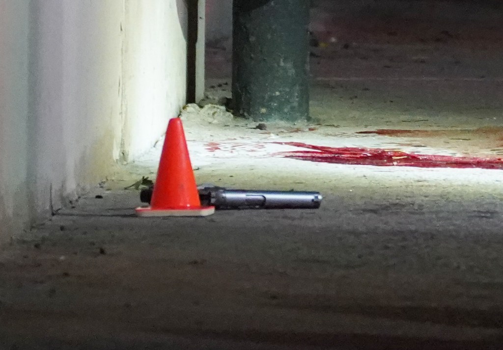 A gun and an orange traffic cone lie on the ground next to a large pool of blood.