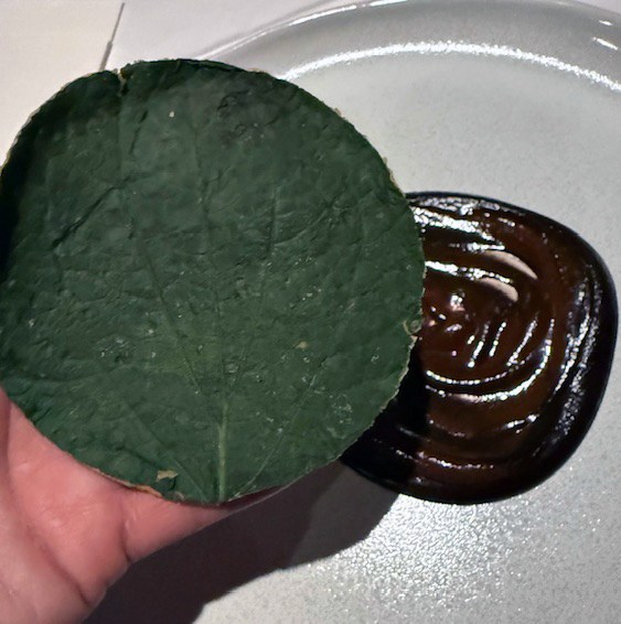 Someone holding up a round green leaf in front of a plate of brown sauce.
