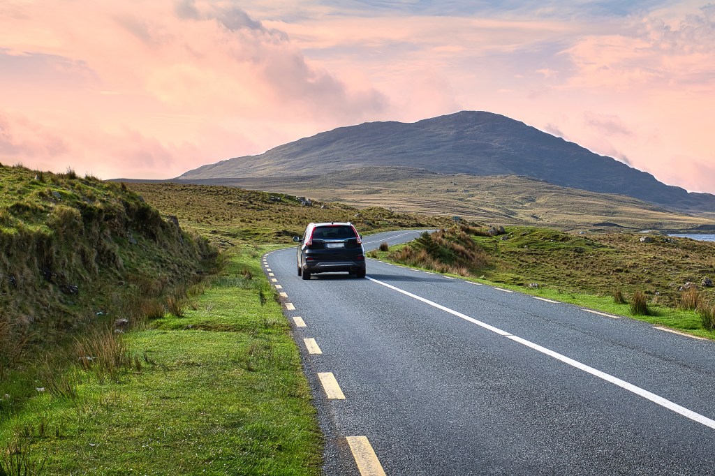 A black car drives on a winding road through a mountainous, green landscape under a pink and orange sky.