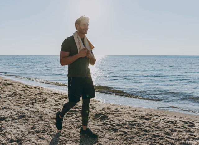 Full size happy young strong athletic toned fit sportsman man in sports clothes towel warm up training walking look aside at sunrise sun dawn over sea sand beach outdoor seaside in summer day morning.