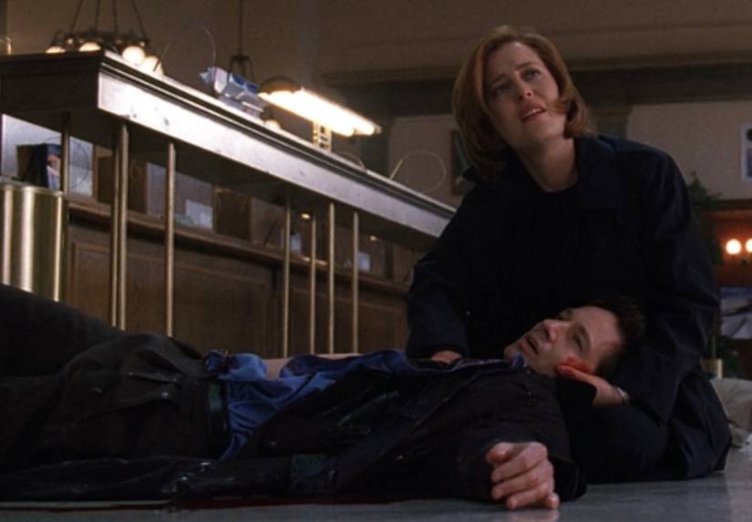 Dana Scully (Gilian Anderson) sits on the floor of a bank cradling the bleeding Fox Mulder (David Duchovny) in The X-Files episode "Monday"
