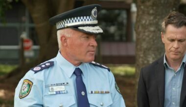 A police officer wearing a NSW Police uniform and cap looks to his left. Standing next to him is a man in a dark jacket and light blue shirt.