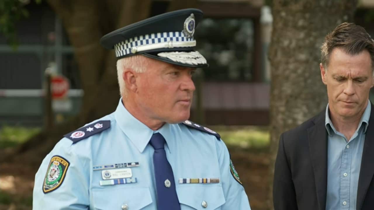 A police officer wearing a NSW Police uniform and cap looks to his left. Standing next to him is a man in a dark jacket and light blue shirt.