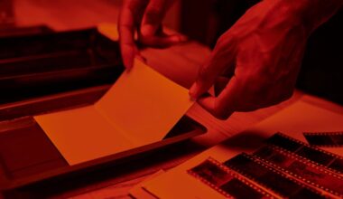 A person's hands develop a photo in a darkroom, holding photographic paper over a tray of liquid. Strips of film negatives and other photographic materials are visible on the wooden table under red lighting.