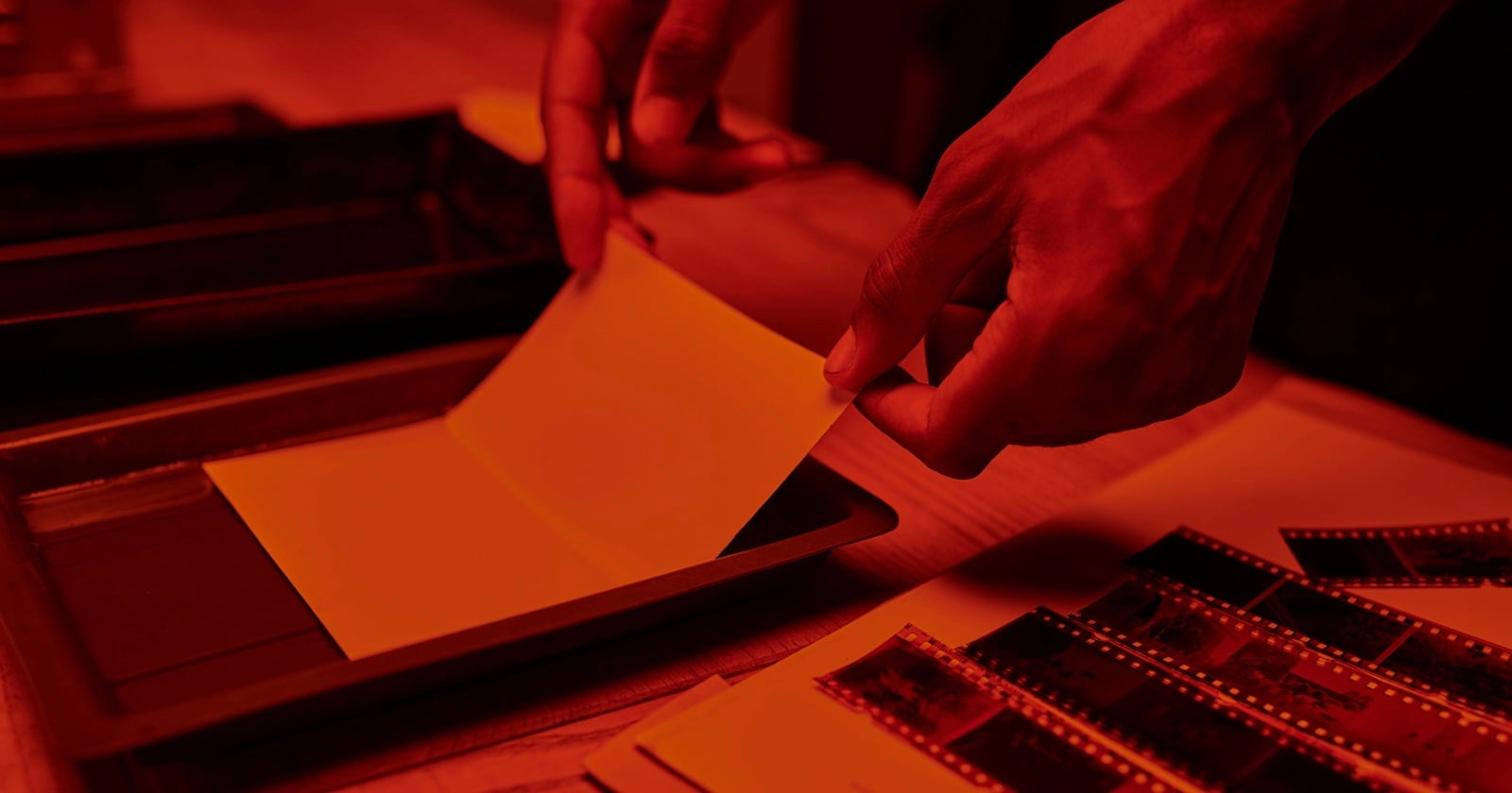 A person's hands develop a photo in a darkroom, holding photographic paper over a tray of liquid. Strips of film negatives and other photographic materials are visible on the wooden table under red lighting.