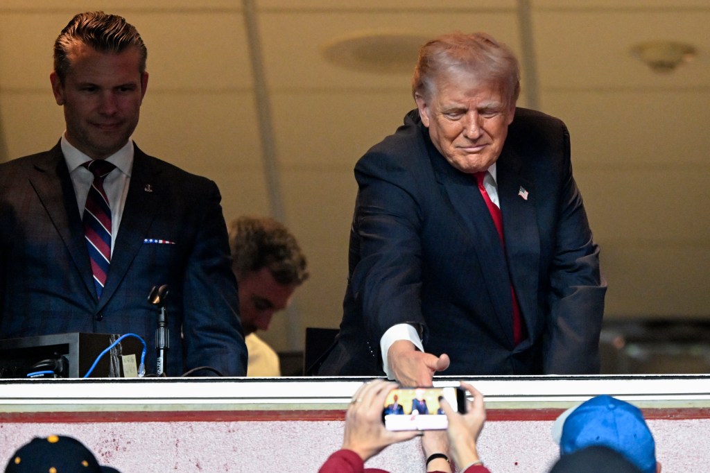 Donald Trump reaches out to shake hands with a fan while attending a football game, with Pete Hegseth looking on.