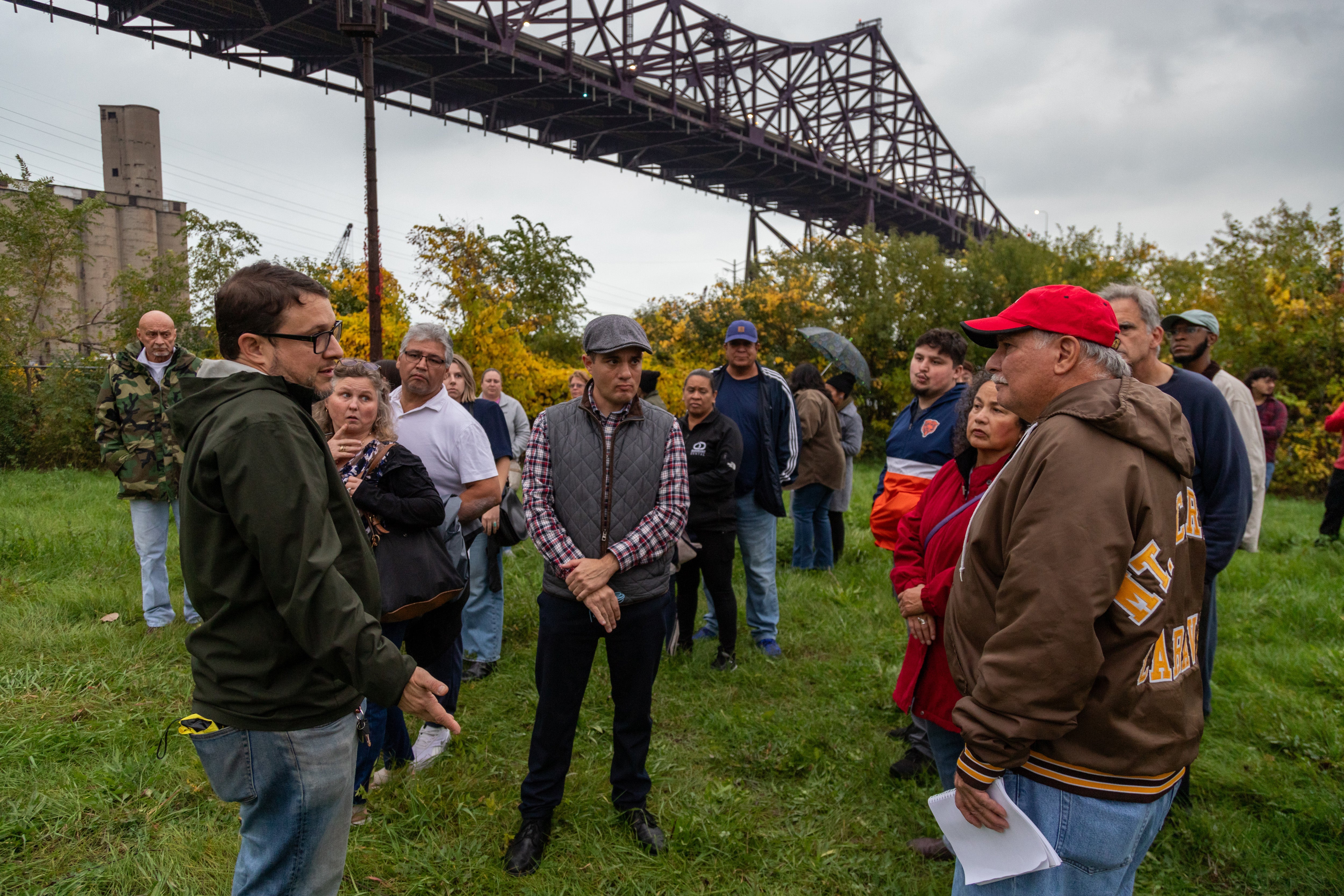 people stand and talk in a field under a bridge
