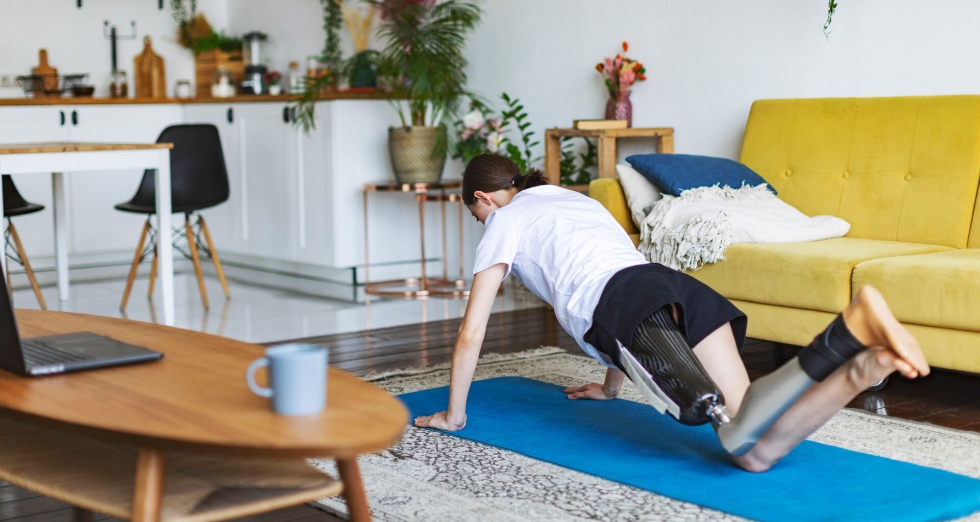 person facing away from the camera with a prothetic leg performing a push up from their knees on a blue exercise mat in a living room setting with a yellow sofa behind them.
