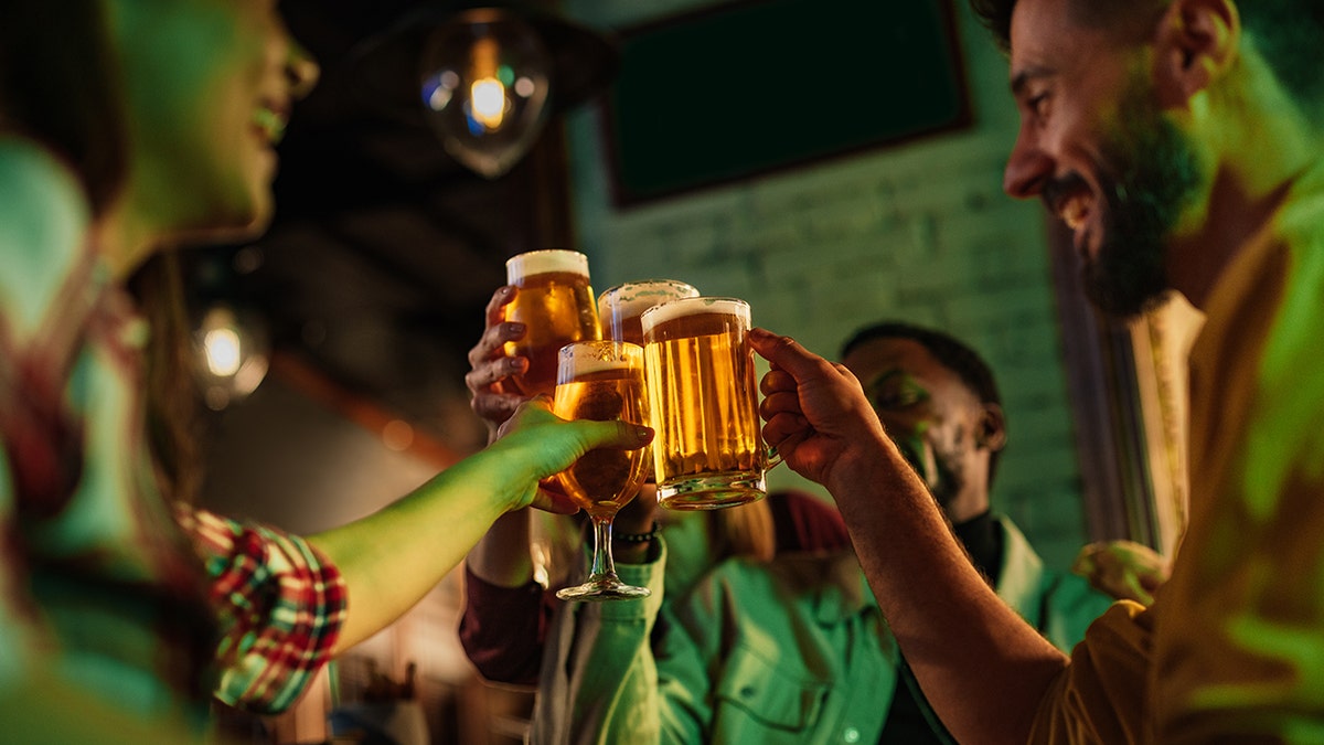 Group of young people celebrating and toasting with beers at a bar.