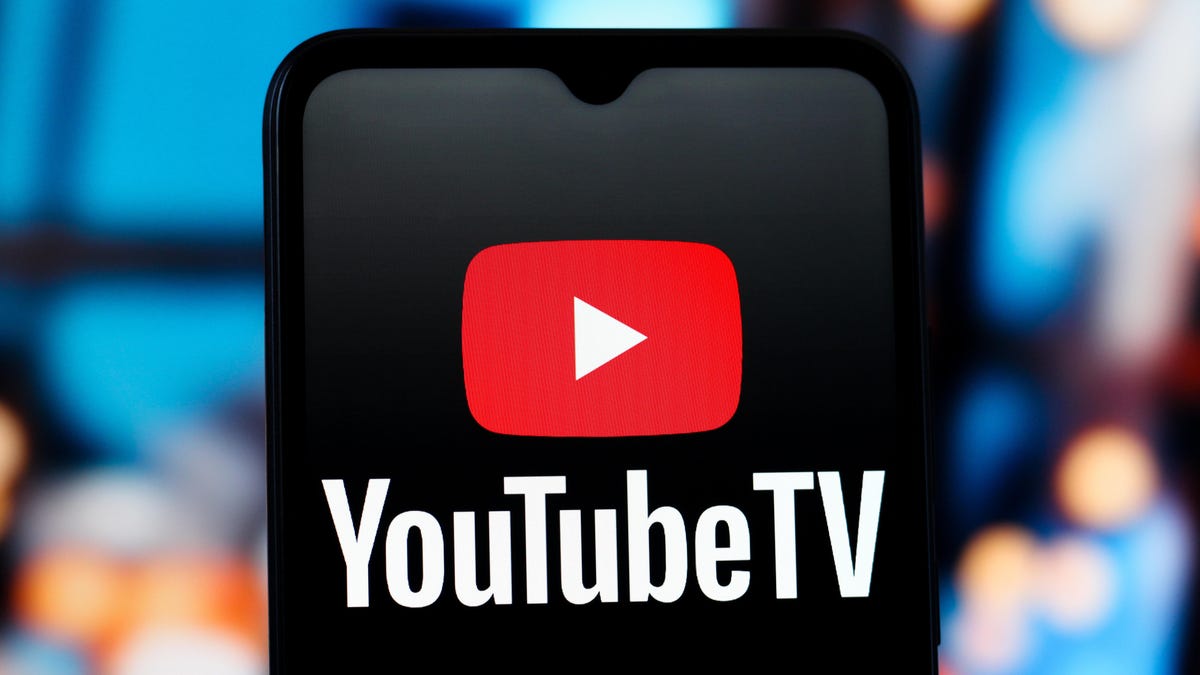 the YouTube TV logo on a black mobile phone in front of a blurry set of monitors
