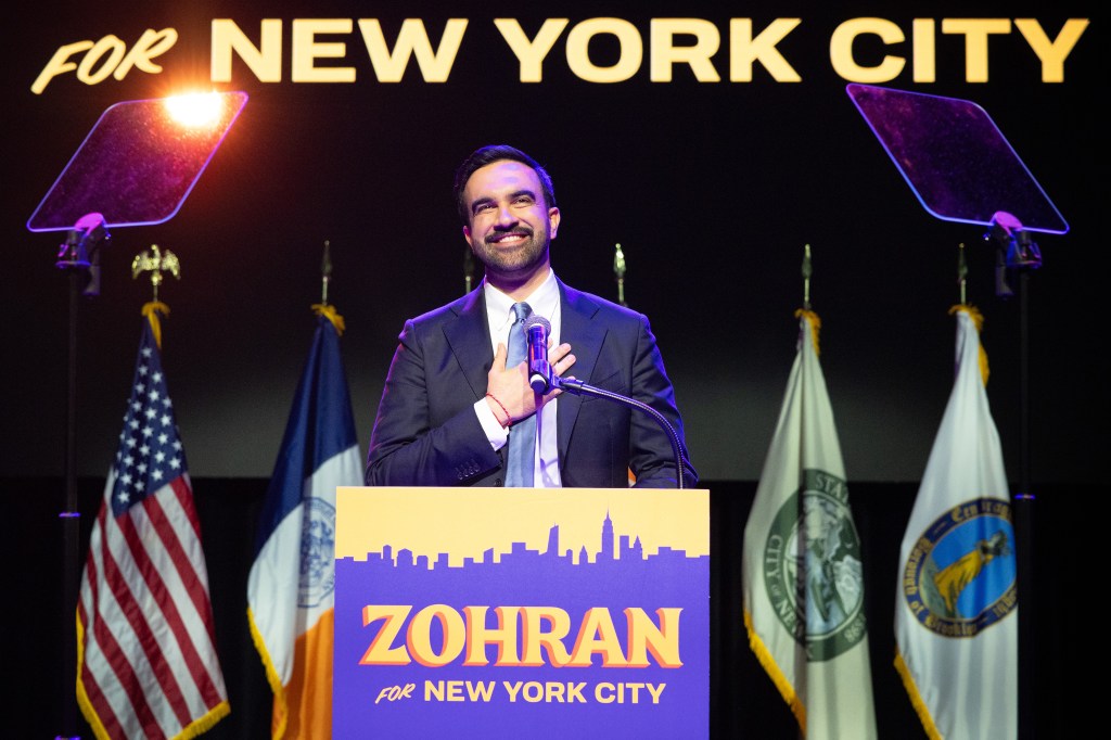 Zohran Mamdani, a smiling man in a suit, speaks at a podium with “Zohran for New York City” on the front, flanked by American and New York City flags.