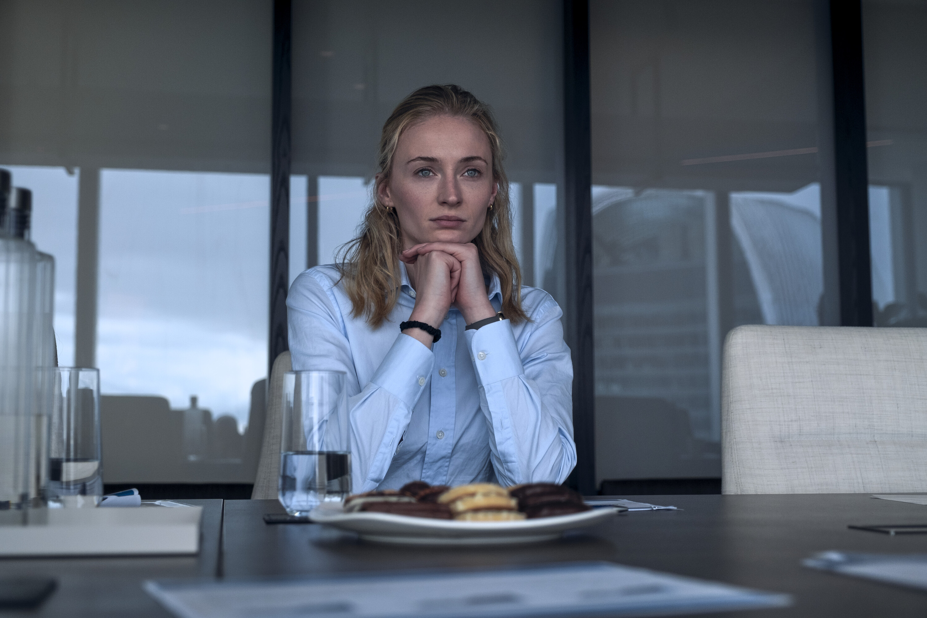 Sophie Turner as Steal, in a blue button-down shirt, sitting at a conference table with her hands clasped under her chin.