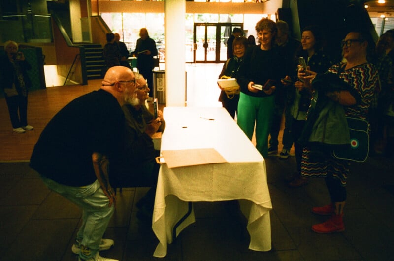 A group of people stand around a table where two men are seated, possibly at a signing event. One man leans in while others smile and take photos. The scene takes place indoors with bright lighting and a window in the background.