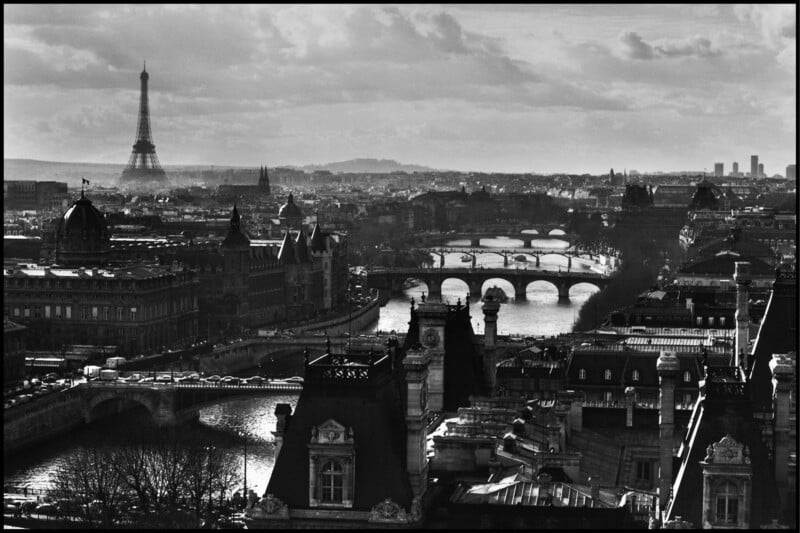 A black-and-white view of Paris featuring the Eiffel Tower in the distance, several bridges crossing the Seine River, and historic city buildings in the foreground under a cloudy sky.