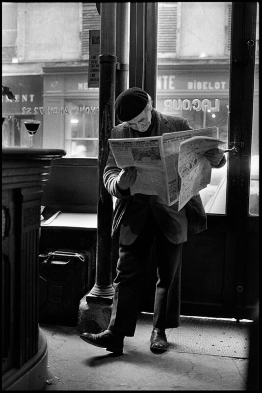 An elderly man wearing a beret stands in a café, leaning against a pole while reading a newspaper. A glass of wine sits on the bar beside him, and the street is visible through the window behind him.