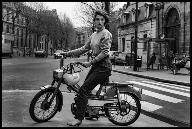 A woman in a sweater and jeans sits on a moped at a city street intersection, looking at the camera. Leafless trees and several people on a bench are visible in the background.