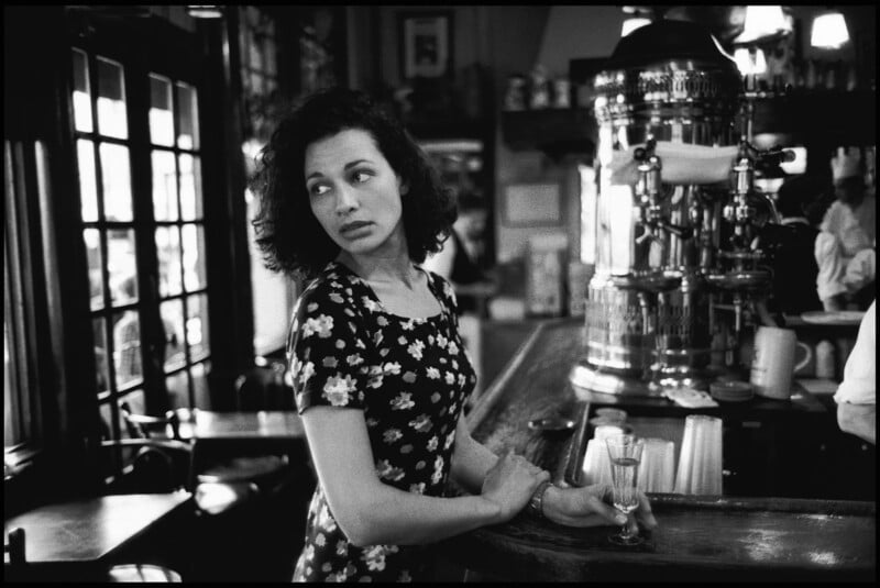 A woman with curly dark hair in a floral dress stands at a bar, looking to her left with a pensive expression. An empty glass sits on the counter, and the background shows a vintage-looking café interior.