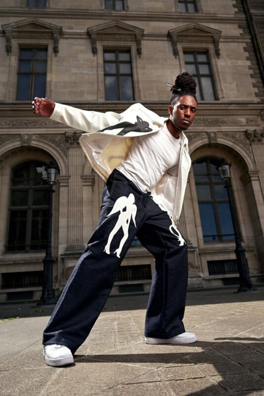 A person posing confidently outdoors in front of a historic building, wearing wide-leg jeans with white silhouette designs, a white T-shirt, a white jacket, and white sneakers.