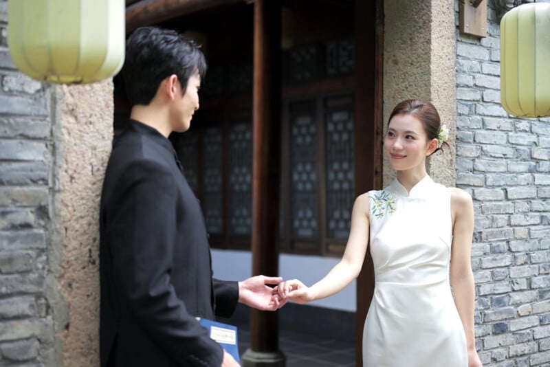 A woman in a white dress and a man in a black suit stand outside a building with gray brick walls, holding hands and smiling at each other. Lanterns hang nearby, and wooden doors are visible in the background.