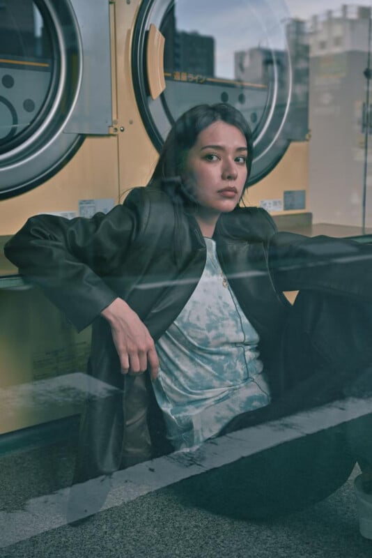 A woman with long dark hair sits on the floor in front of yellow washing machines, looking thoughtfully ahead. She wears a dark jacket and light shirt. The photo is taken through a window, adding reflections.