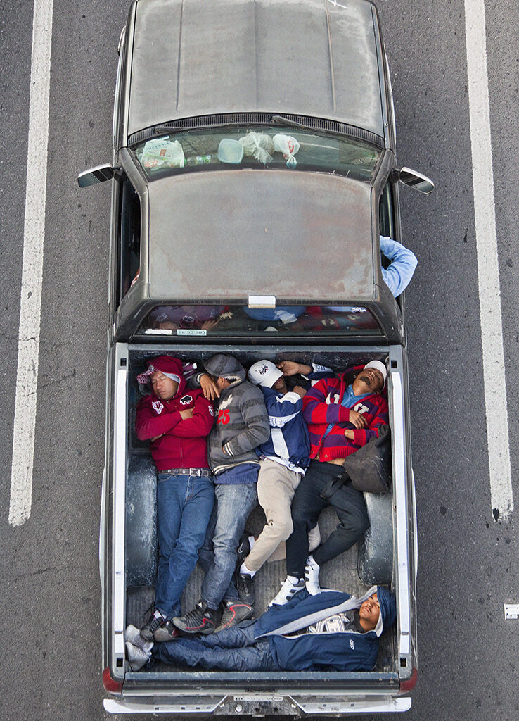 Aerial view of six people lying closely together in the bed of a pickup truck parked between two white lines on a road.