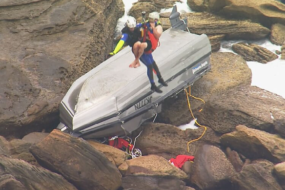 A man is lifted from the rocks to a rescue helicopter, near the flipped boat.
